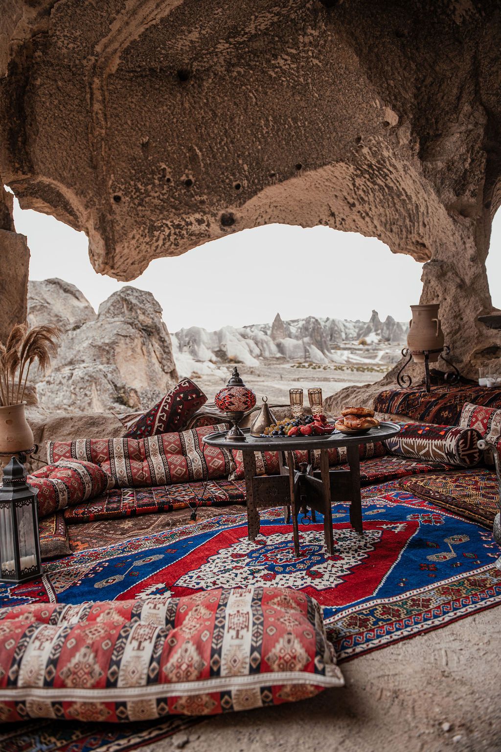 A cozy cave dwelling in Cappadocia with colorful Turkish rugs, cushions, and a small table overlooking rocky landscape.