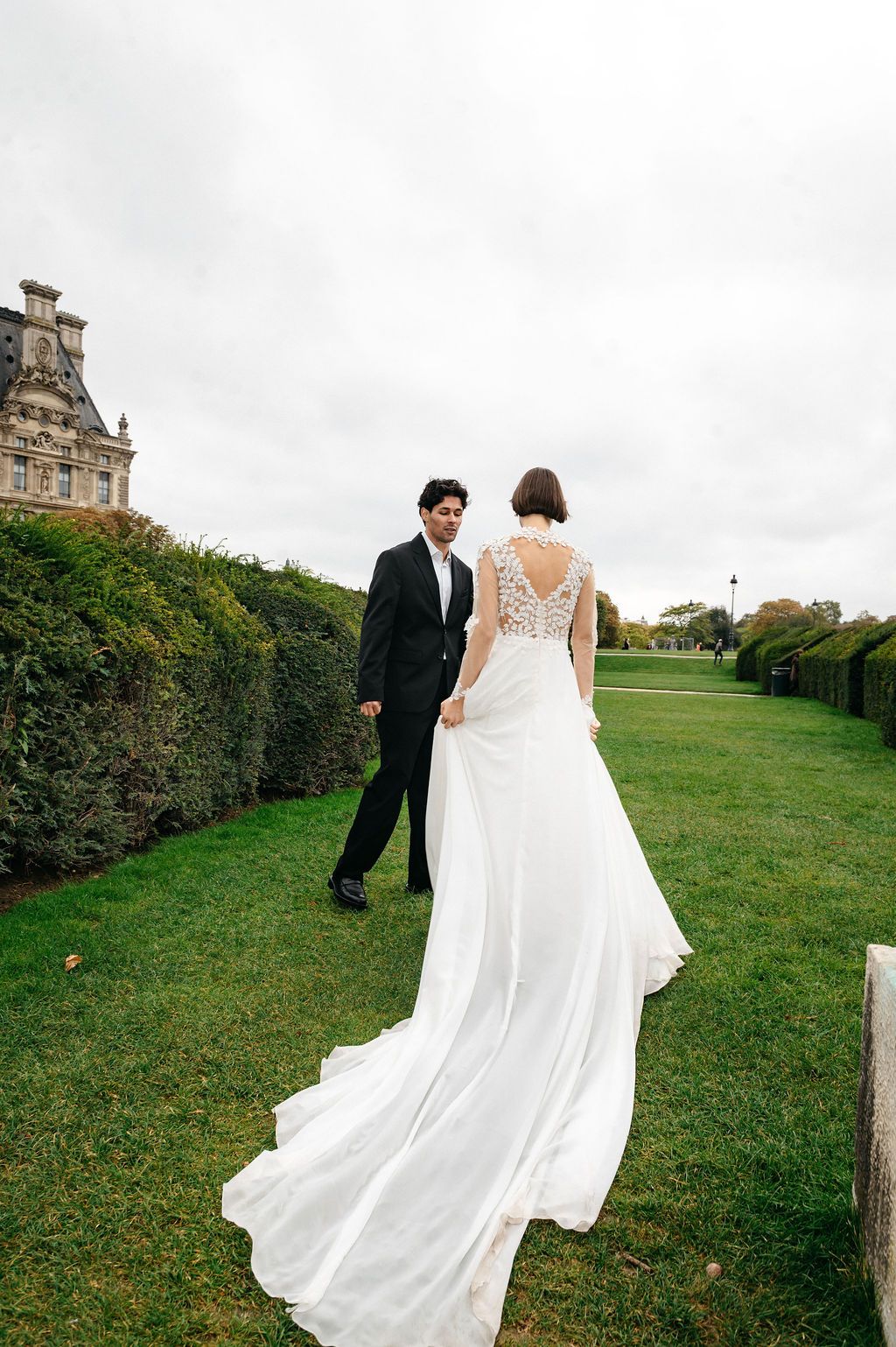 Bride in white lace gown with train walks with groom in black suit through lush garden path near historic building.