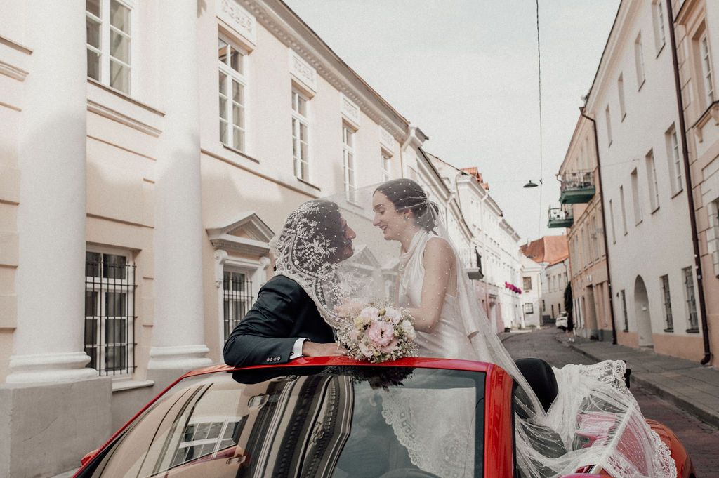Bride and groom sharing a romantic kiss standing through the sunroof of a red convertible car on a European street.