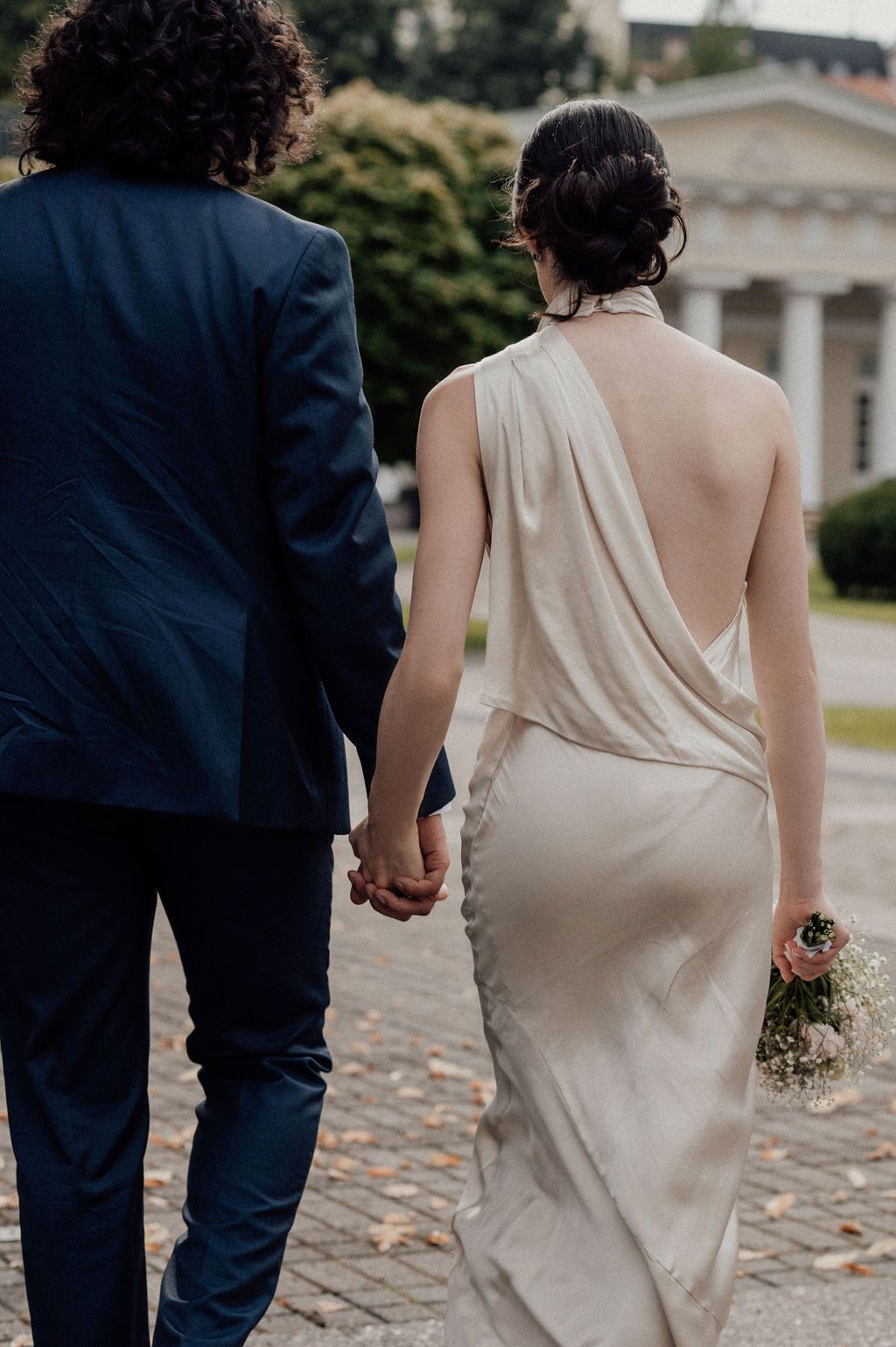 A bride in a backless ivory gown holds hands with a groom in a navy suit while walking on a cobblestone path.