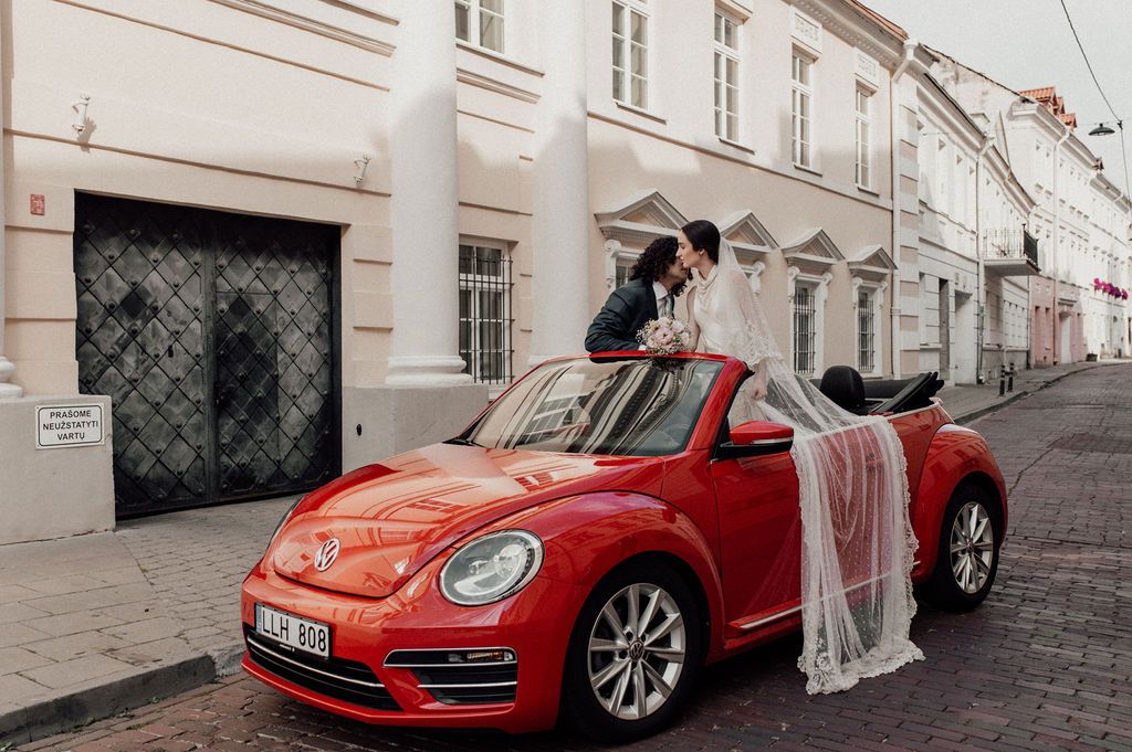 Bride and groom kissing on top of a red Volkswagen Beetle on a cobblestone street in a European city.