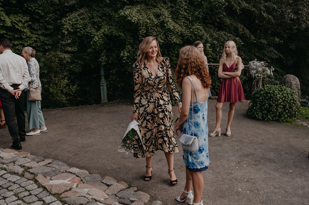 Guests in elegant dresses socializing outdoors at a wedding reception on a stone patio surrounded by lush greenery.