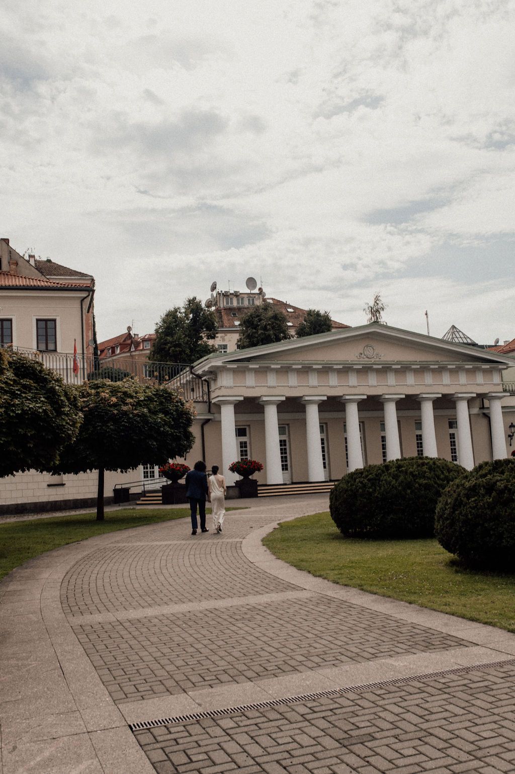 Two people stroll along a brick pathway toward a neoclassical building with white columns in a lush European city square.