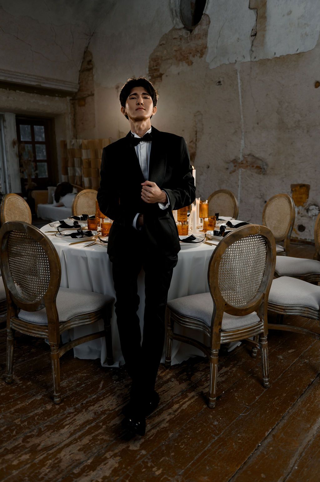 Elegant man in black tuxedo standing confidently beside a formally set dining table with gold accents.