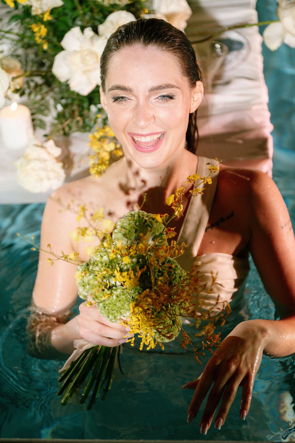 Smiling woman holding yellow flowers while relaxing in a pool surrounded by white floral arrangements.