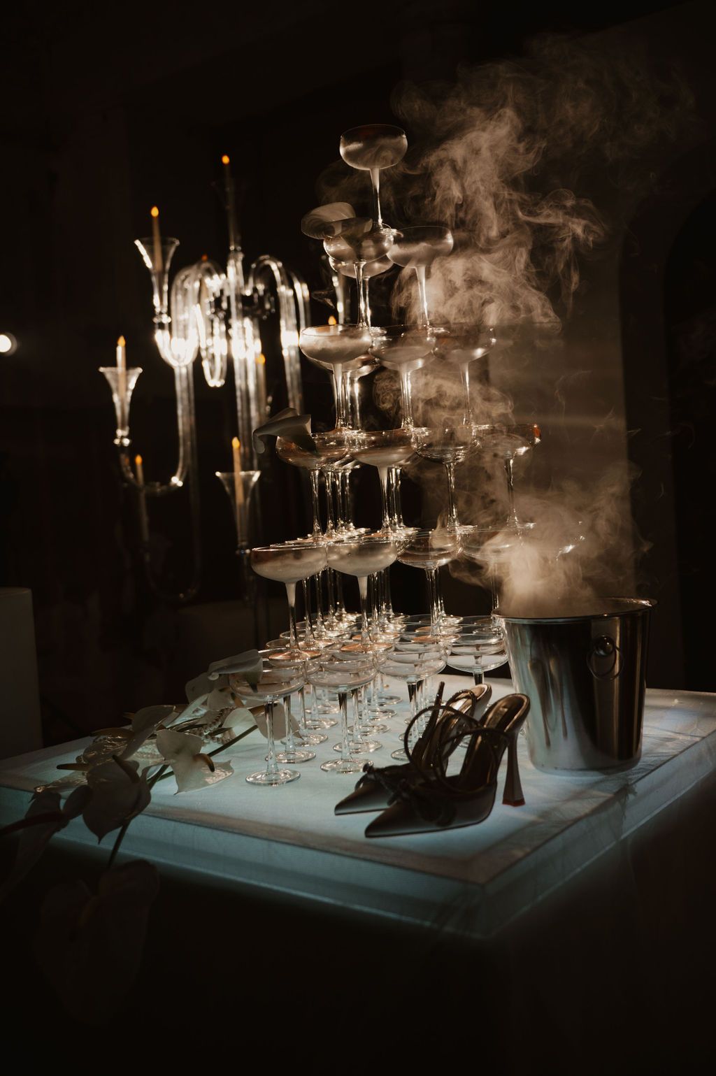 Elegant champagne tower with smoke effect beside high heel shoes on illuminated display table in dark setting.