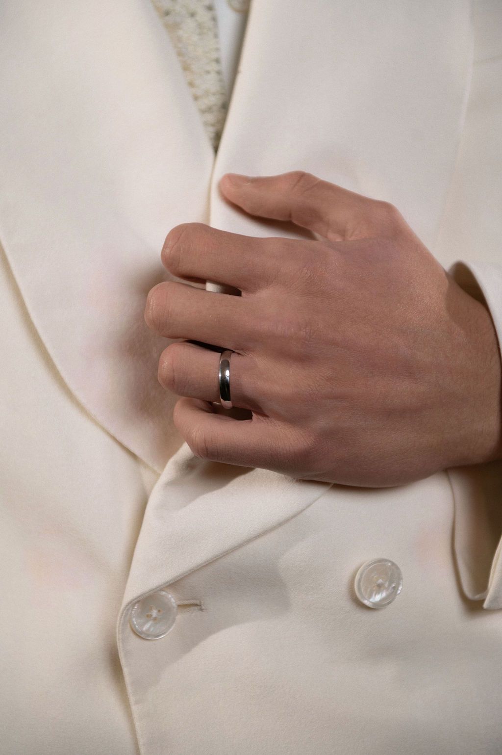 A hand wearing a silver wedding band rests on a white double-breasted blazer with pearl buttons.