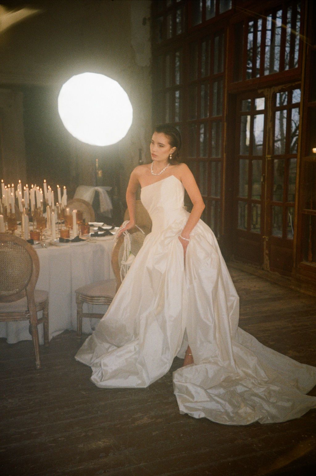 Bride in strapless white ball gown wedding dress poses near candlelit table in elegant rustic venue with glowing orb light.