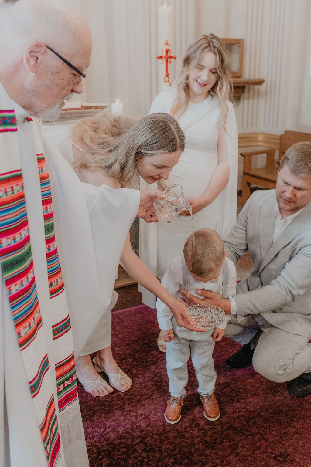 Priest baptizes toddler boy as family gathers around the altar in a church ceremony.