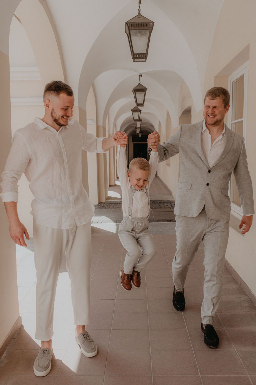 Two men in light suits swing a happy child between them in an elegant arched corridor.