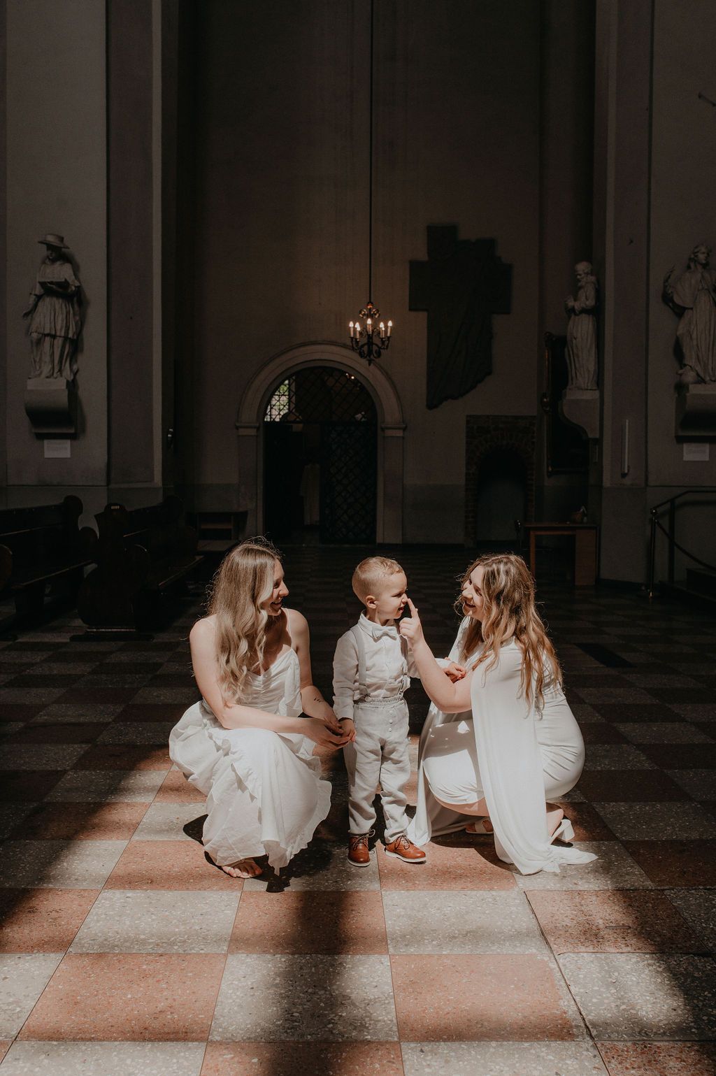 Two brides in white dresses kneel beside a young boy in a suit on a checkered floor inside an elegant church.