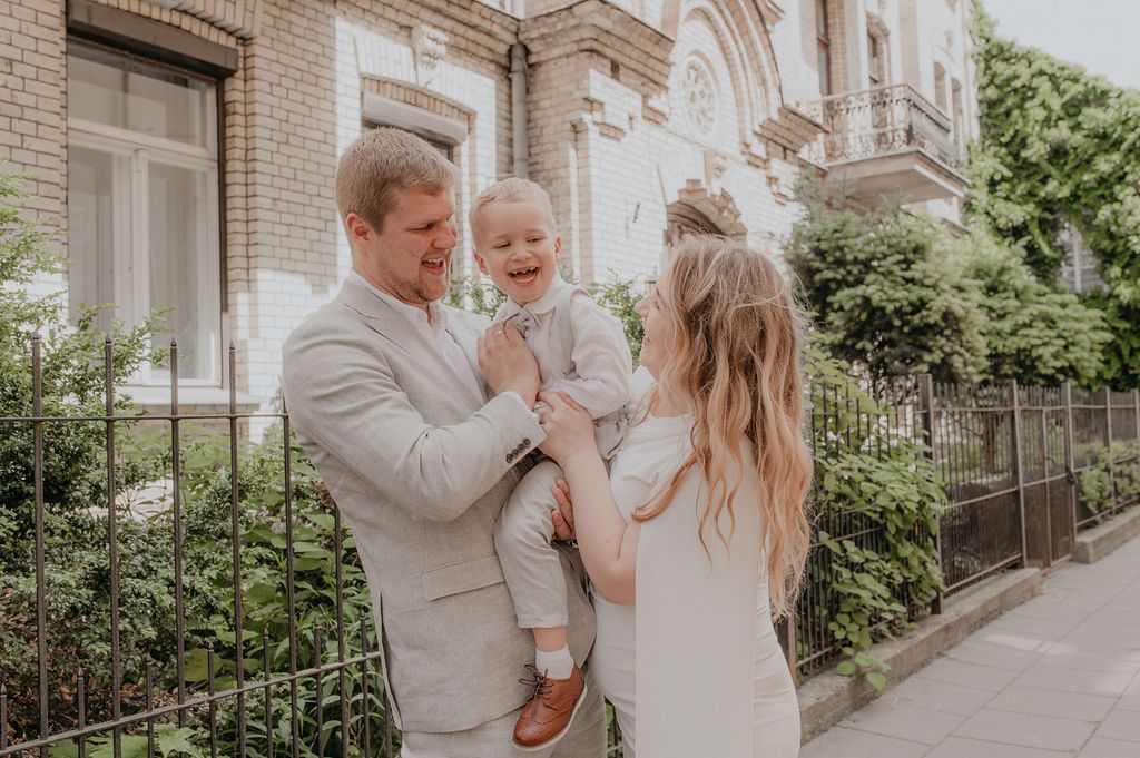Happy family with parents holding laughing toddler boy outdoors near brick building on sunny day.