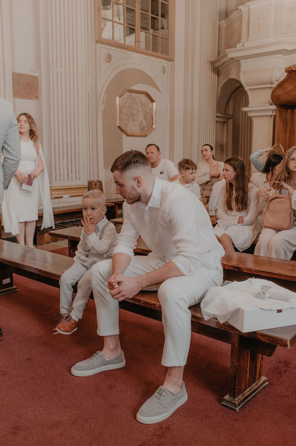 Man and boy in white outfits sit on church pew during a formal ceremony with guests seated in background.