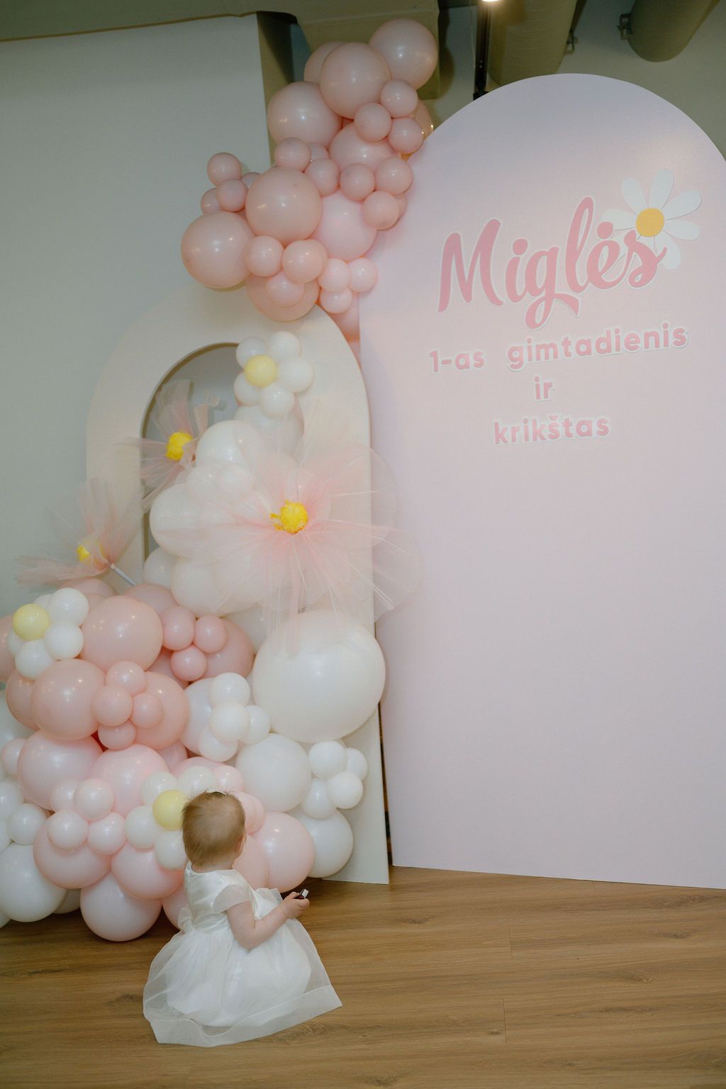 Baby girl in white dress at Miglės 1st birthday and baptism party with pink balloon arch decoration.