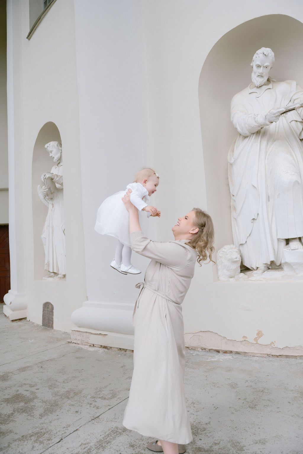 Woman in beige dress lifting baby girl in white dress outdoors near classical white statues.