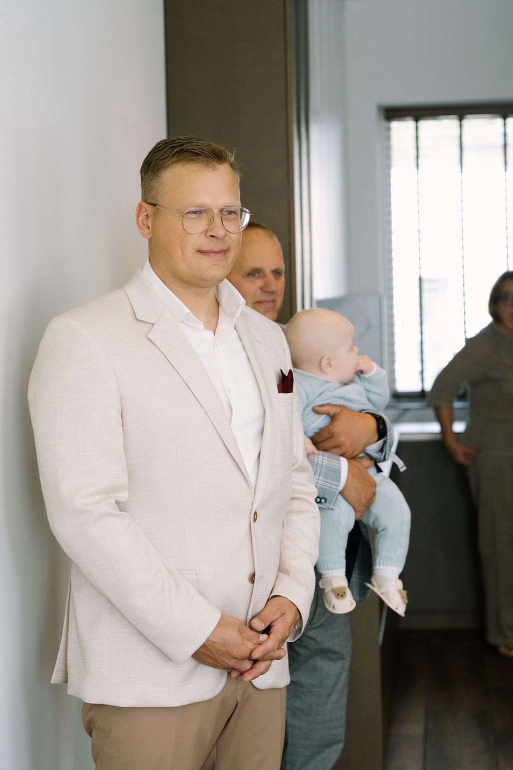 A smiling groom in a beige suit with a burgundy boutonniere stands indoors as a man holds a baby in the background.
