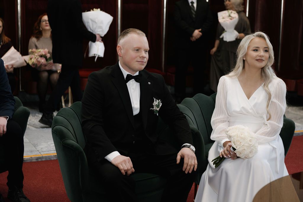 Elegant wedding couple seated at ceremony, groom in tuxedo and bride in white dress holding bouquet.