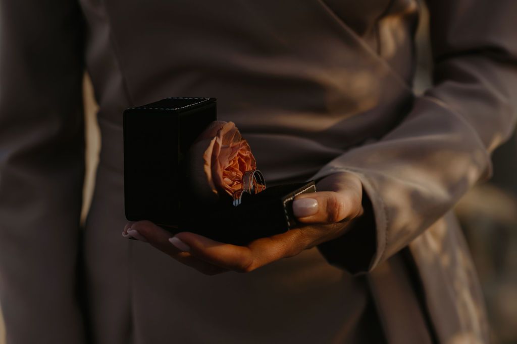 Person in grey satin holding open black velvet box with a pink rose inside.