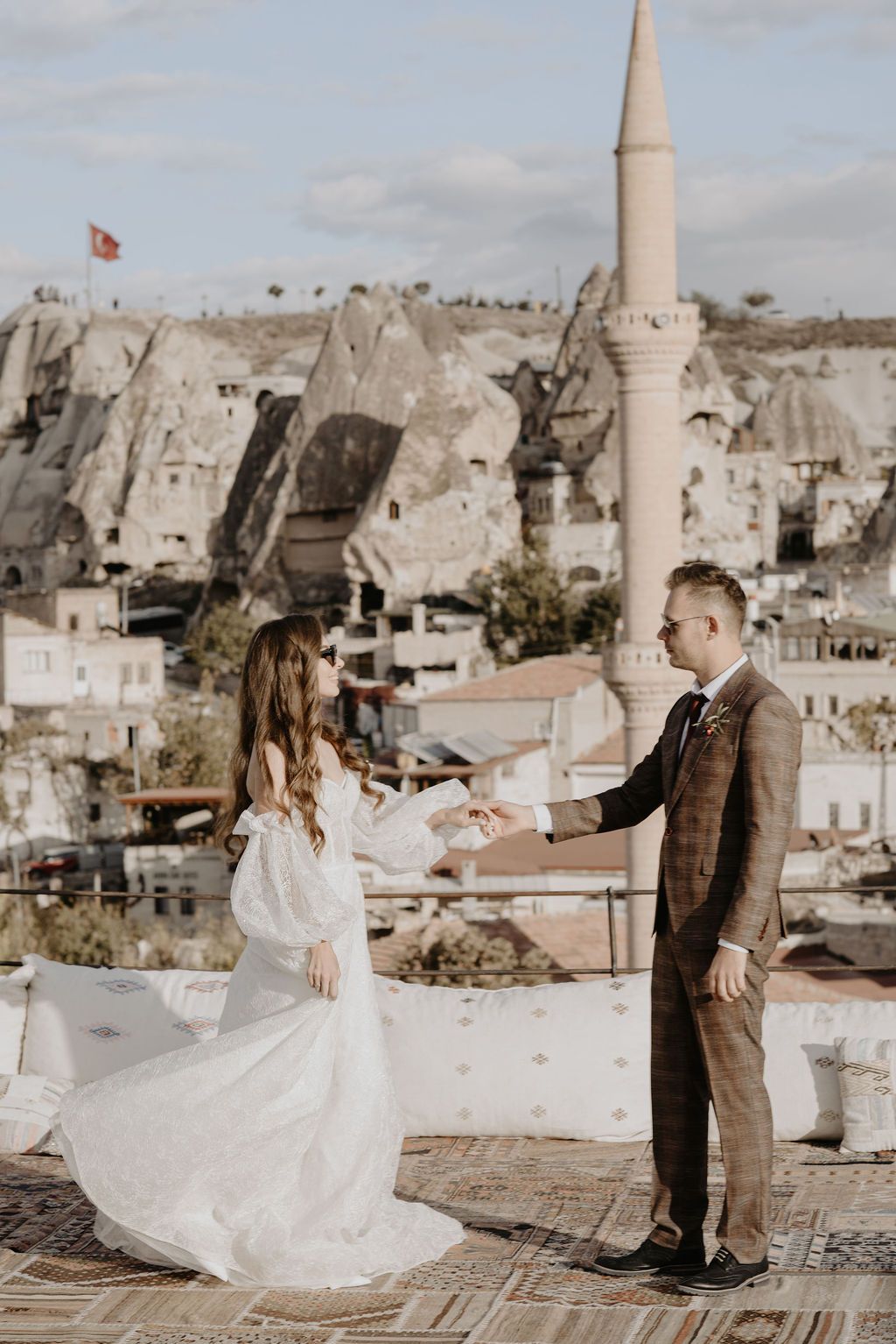 Bride and groom hold hands on a rooftop in Cappadocia, Turkey, with cave dwellings and a mosque minaret in the background.