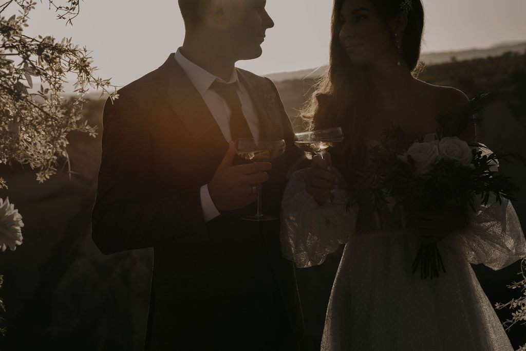 Bride and groom toasting at sunset with wedding bouquet in soft backlit glow.