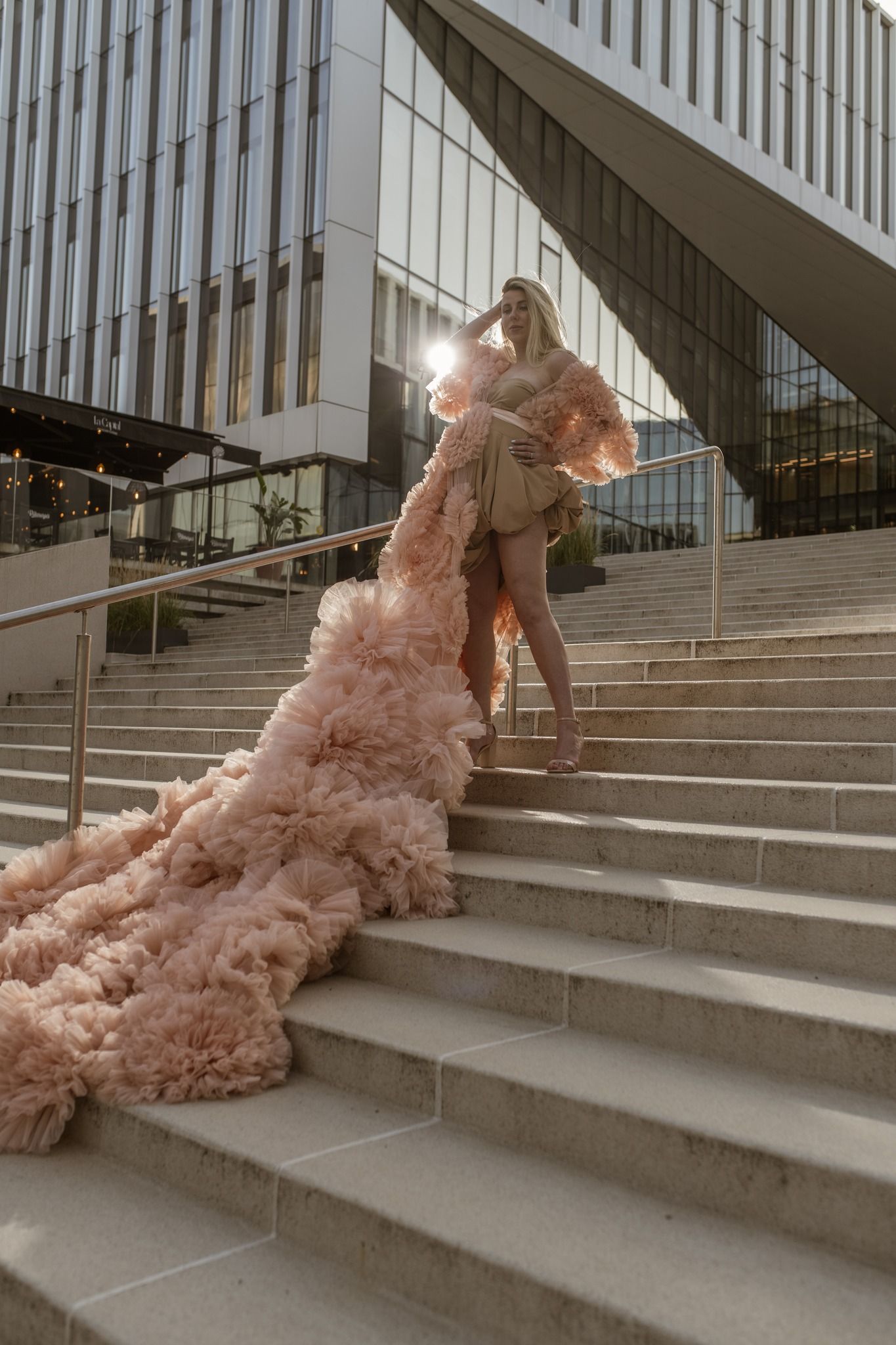 Woman in elegant blush pink ruffle gown with long train poses on marble steps outside modern building.