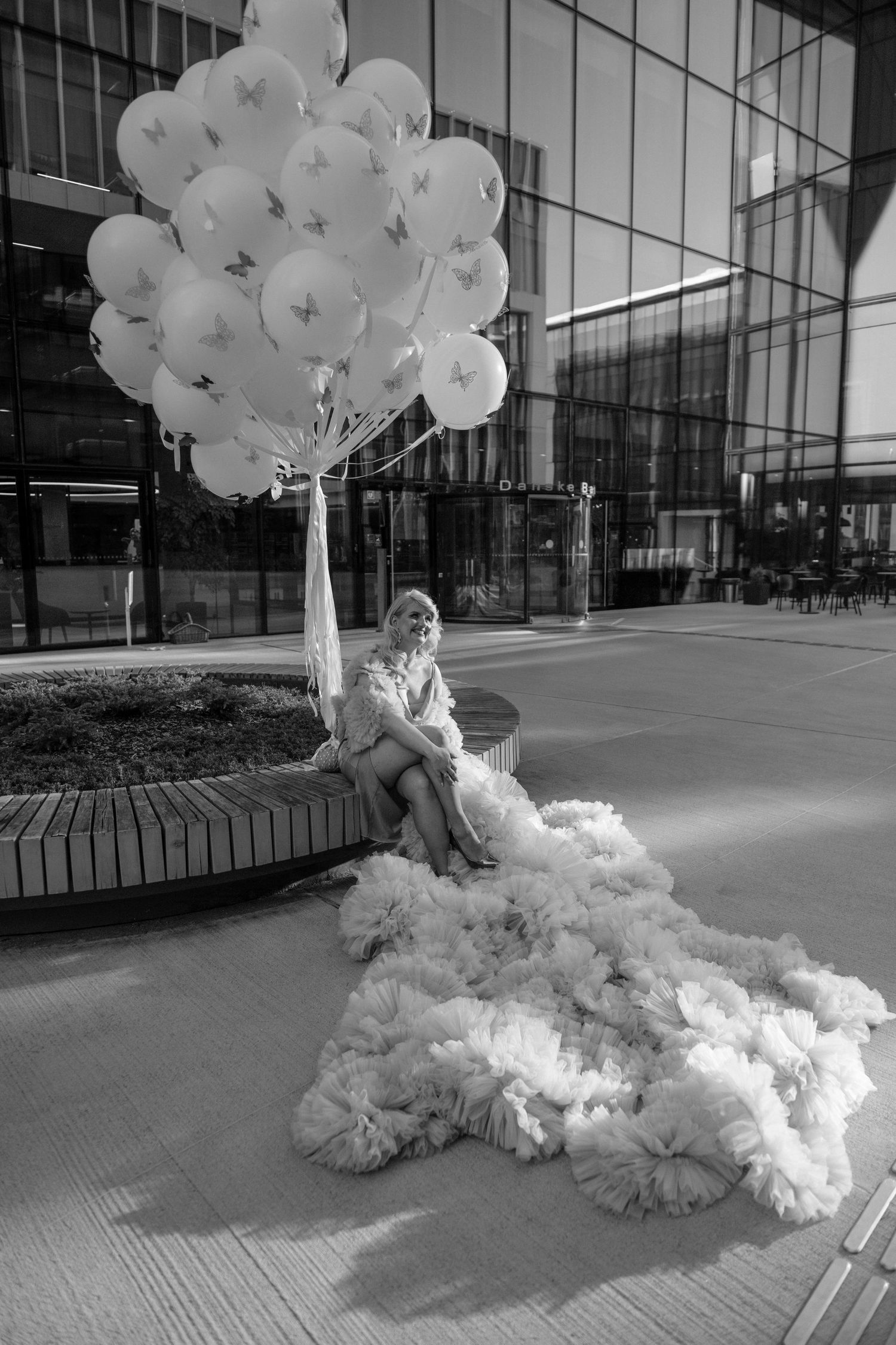 Woman in fluffy white gown holds butterfly balloons outside modern glass building, black and white photo.