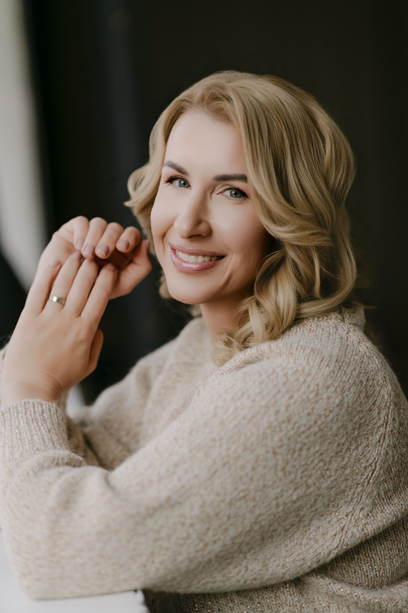Smiling blonde woman in a beige knit sweater posing with hands clasped, wearing a ring, against a dark background.