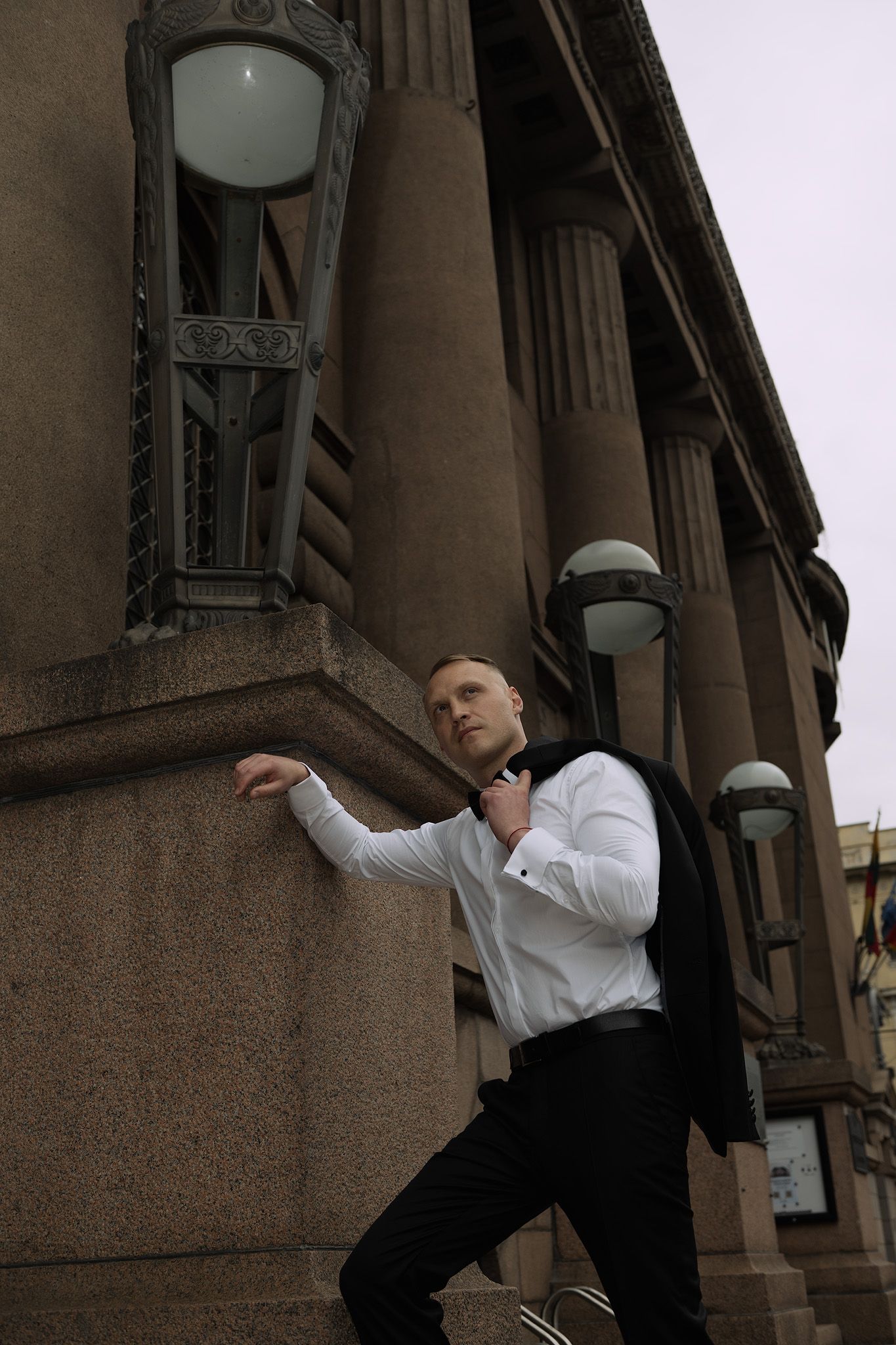 A person in a white shirt and black pants poses dramatically against a classical building with ornate lamp posts.