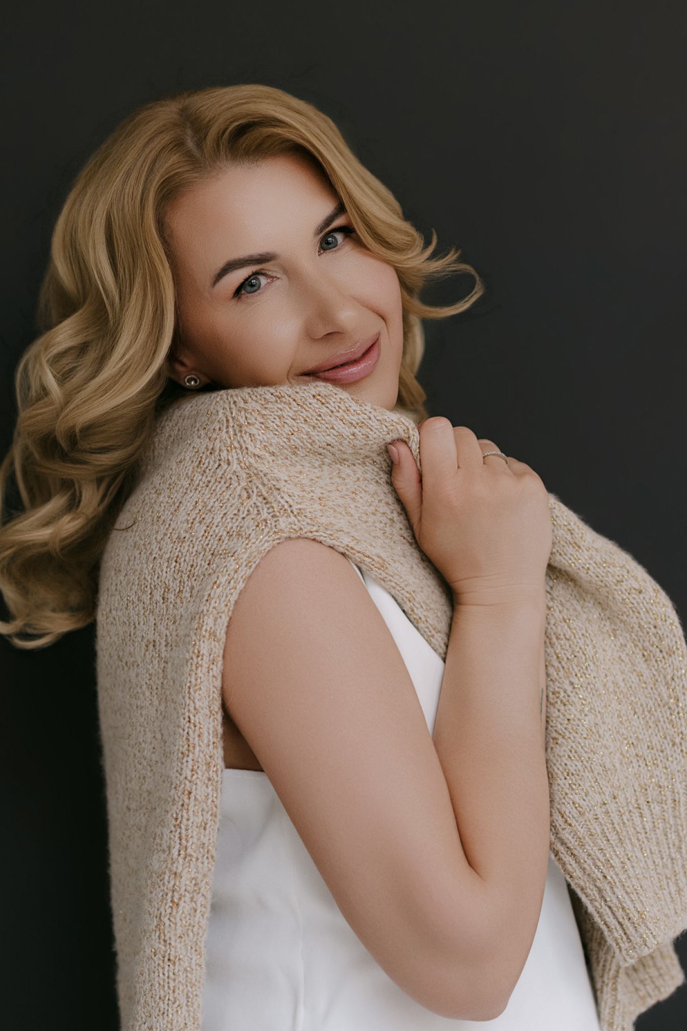 Smiling blonde woman with curly hair holding a beige knit sweater over white top against dark background.