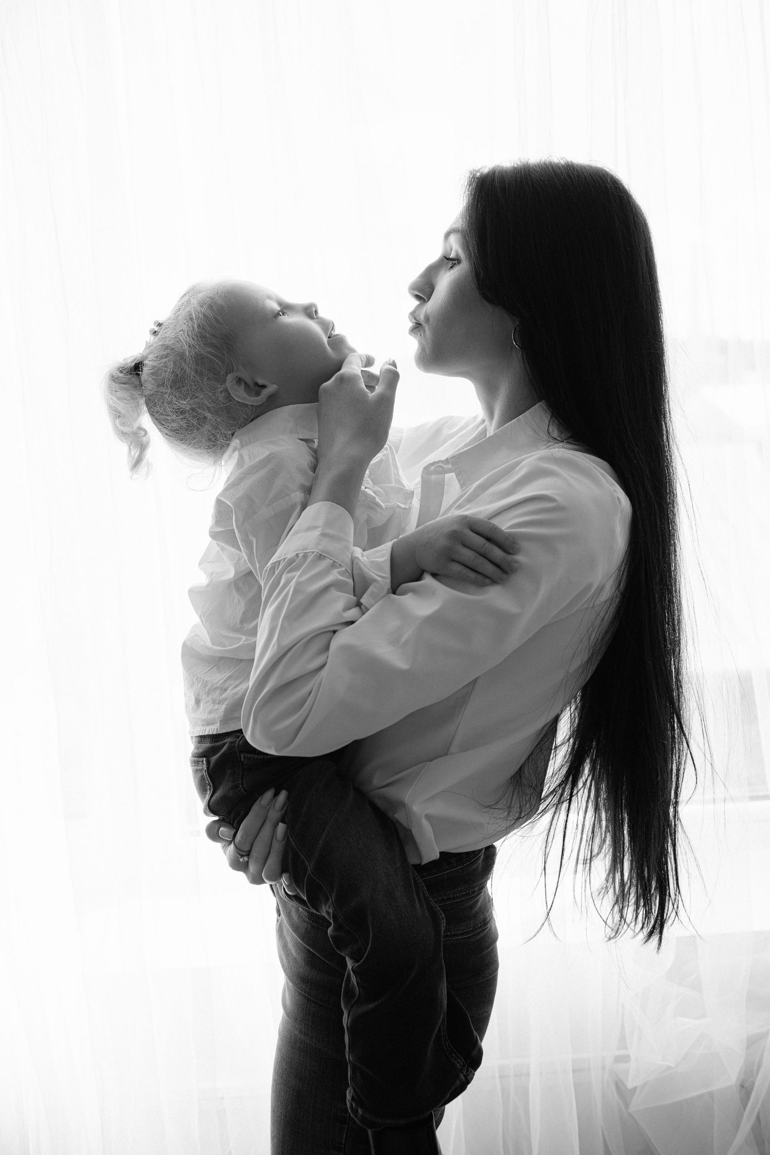 Black and white photo of a mother with long dark hair holding her young blonde toddler near a bright window.