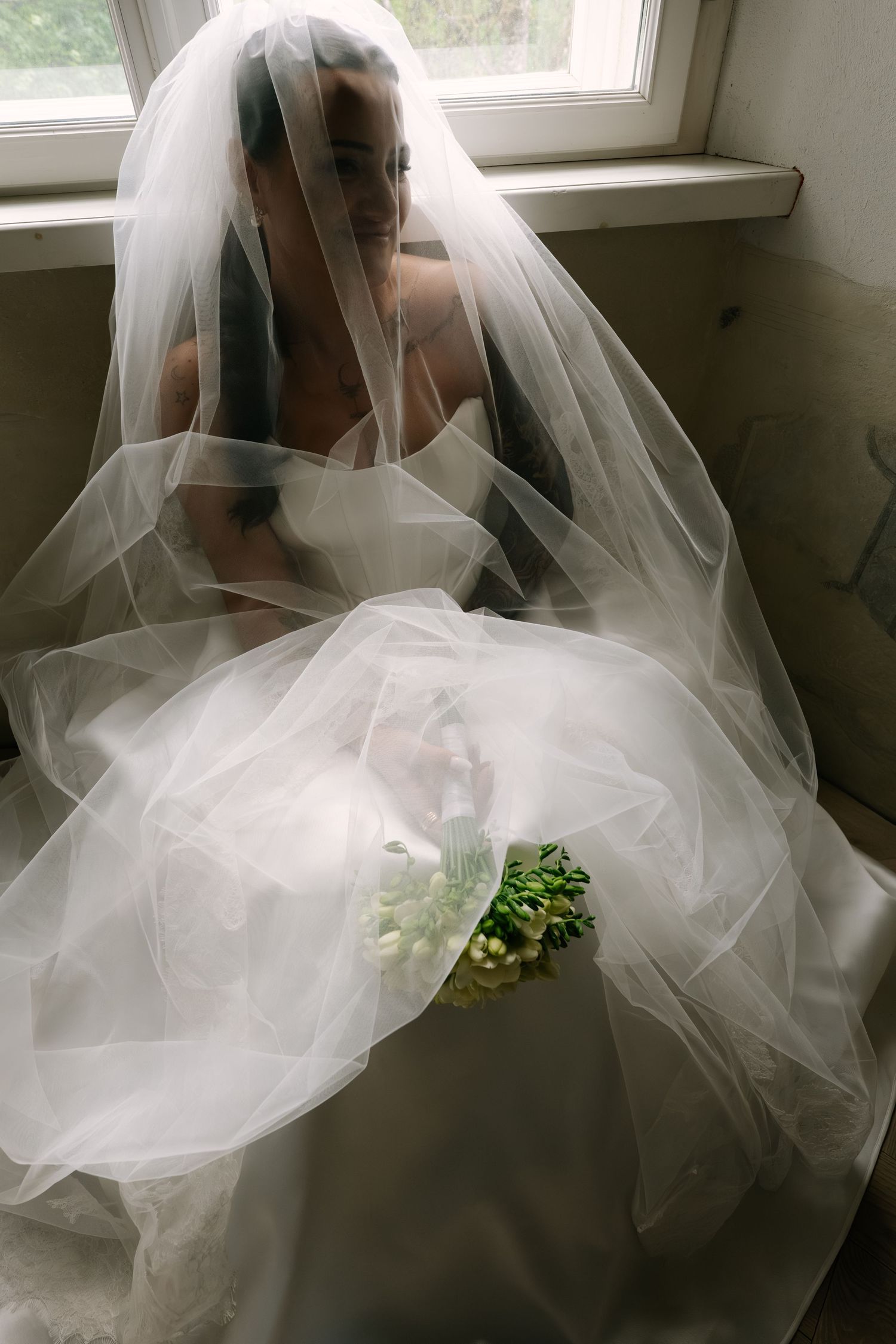 A smiling bride in a sheer tulle veil and strapless white gown sits by a window holding a small bouquet of white and green flowers.