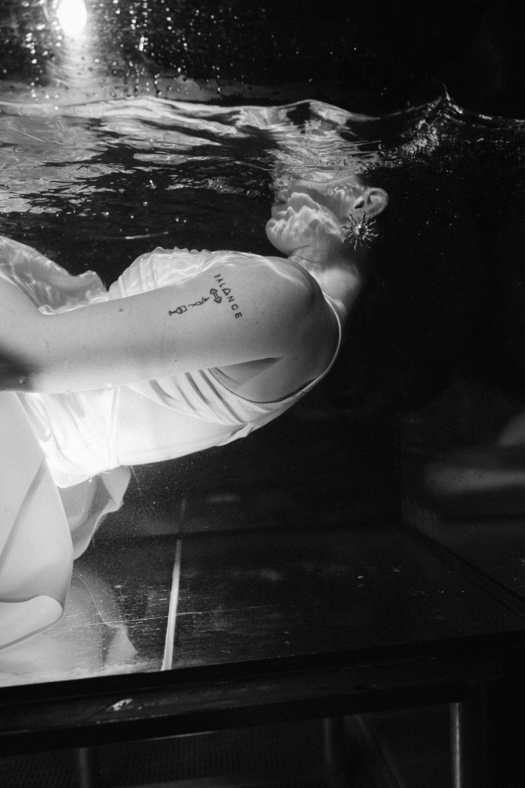 Black and white photo of a woman in a white dress floating underwater beside a glass tank, with a small arm tattoo and star earring.