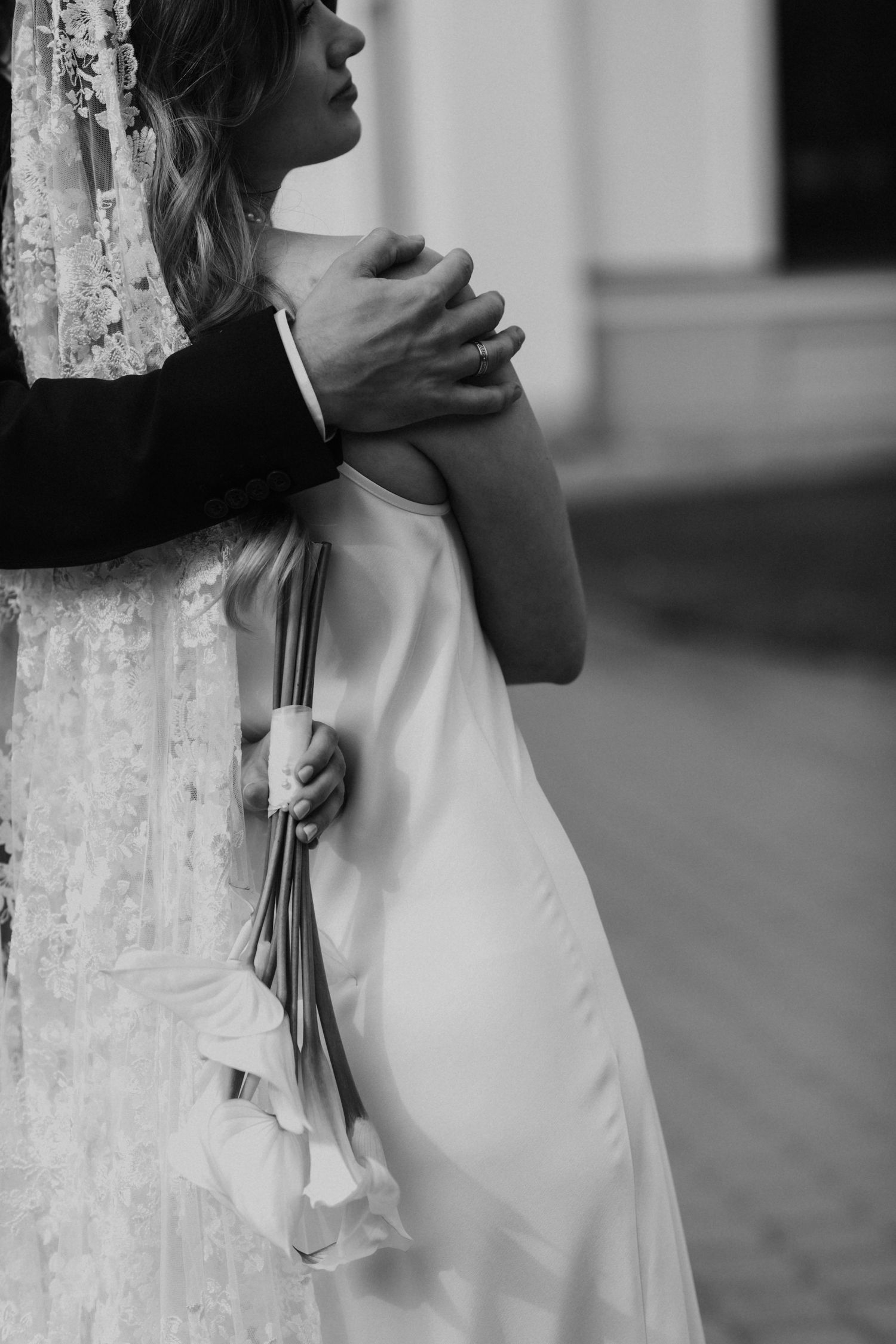 Black and white photo of a bride in a silk slip dress and lace veil holding a calla lily bouquet as a groom embraces her.