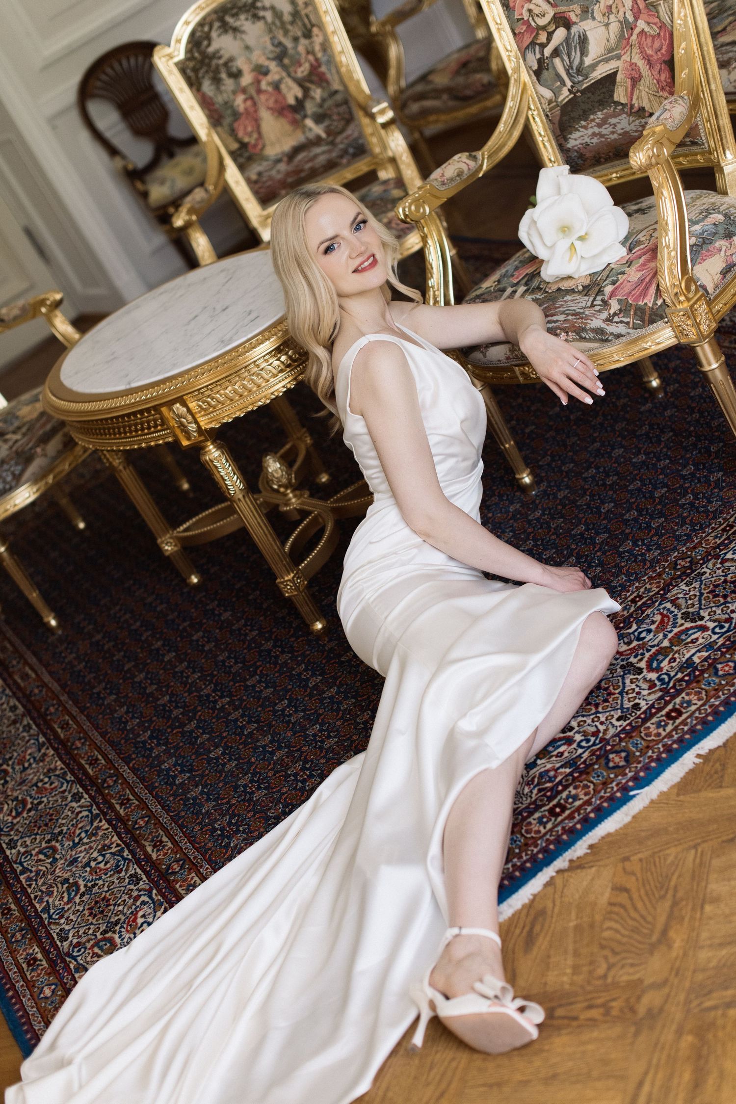 Blonde bride in a white satin gown and heeled sandals poses by a marble table in an ornate room with gilded chairs and a Persian rug.