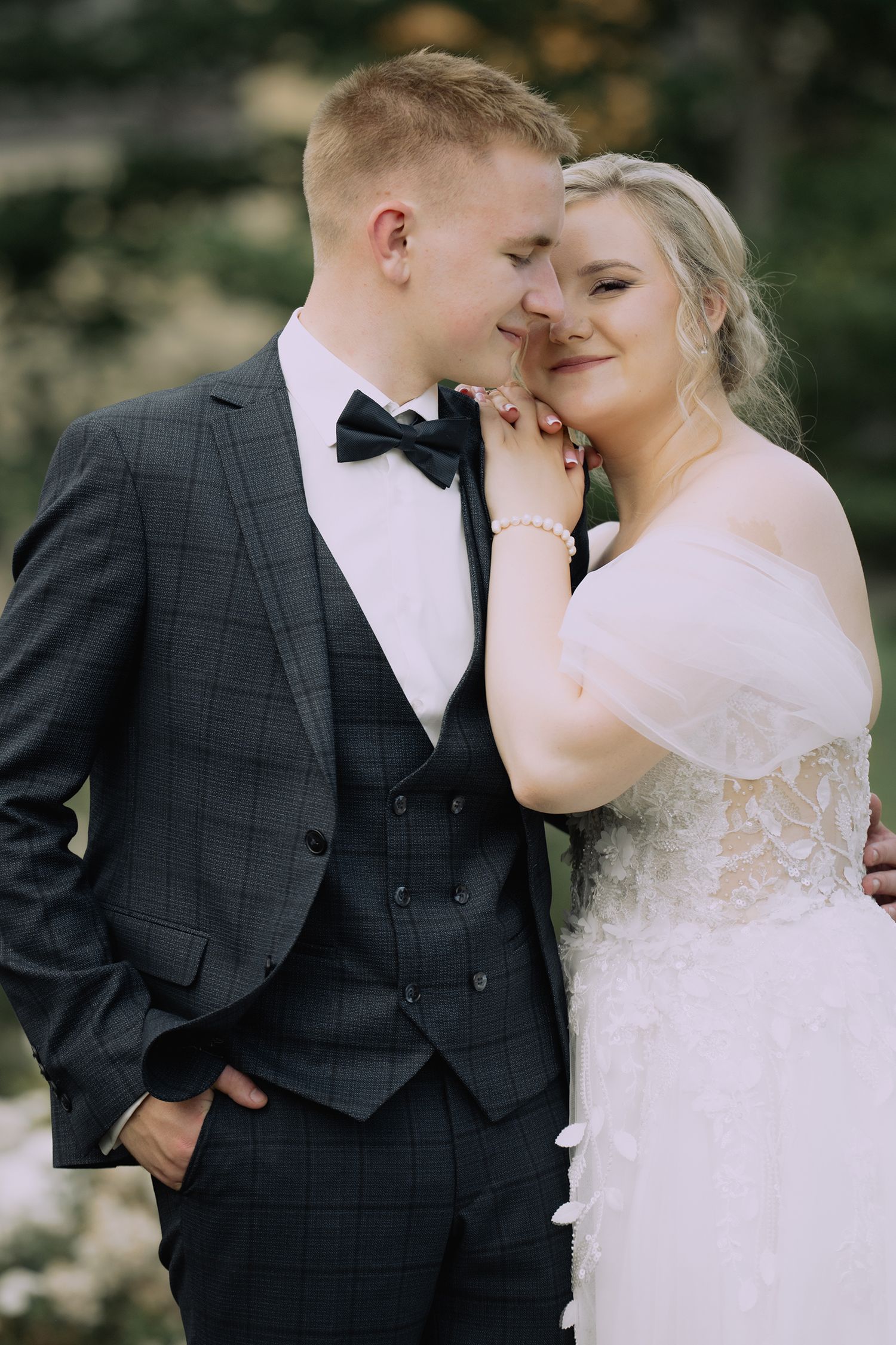 A romantic wedding couple embraces outdoors, with the bride in white lace and groom in charcoal suit and bowtie.