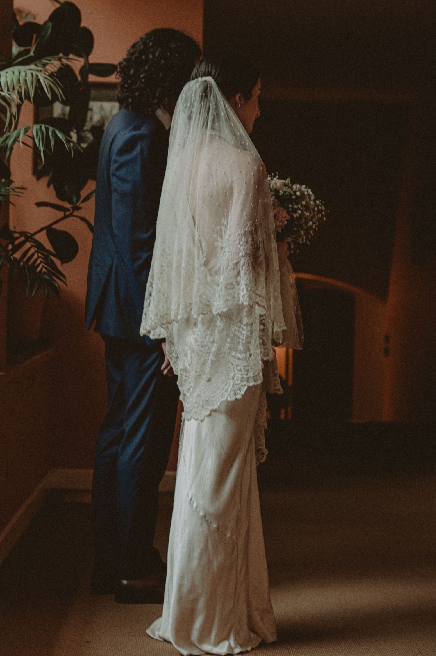 Couple in wedding attire with bride wearing flowing white lace dress and groom in navy suit standing in dim lighting.