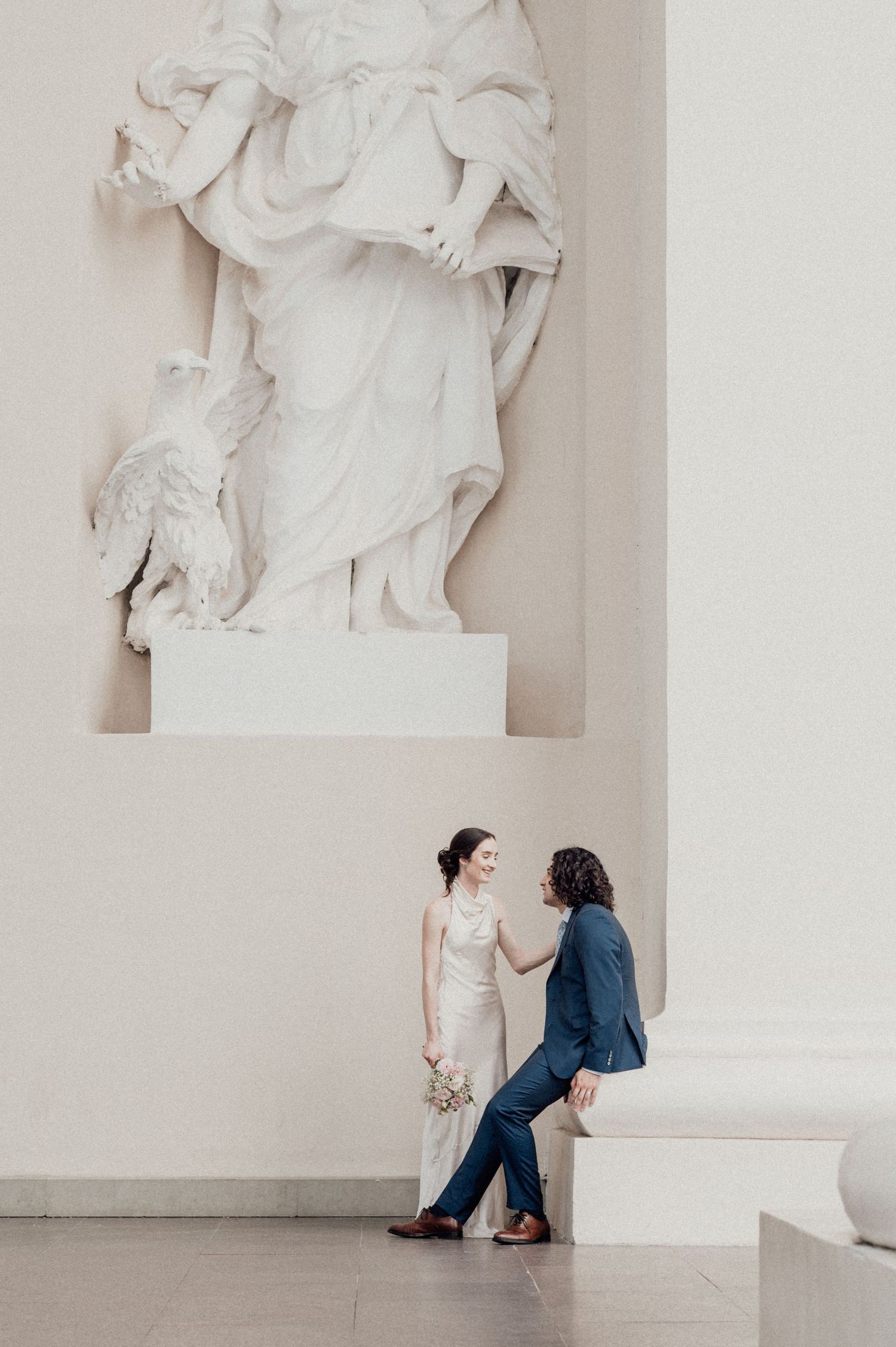 Artistic wedding photo of couple sitting against white wall with classical sculpture above.