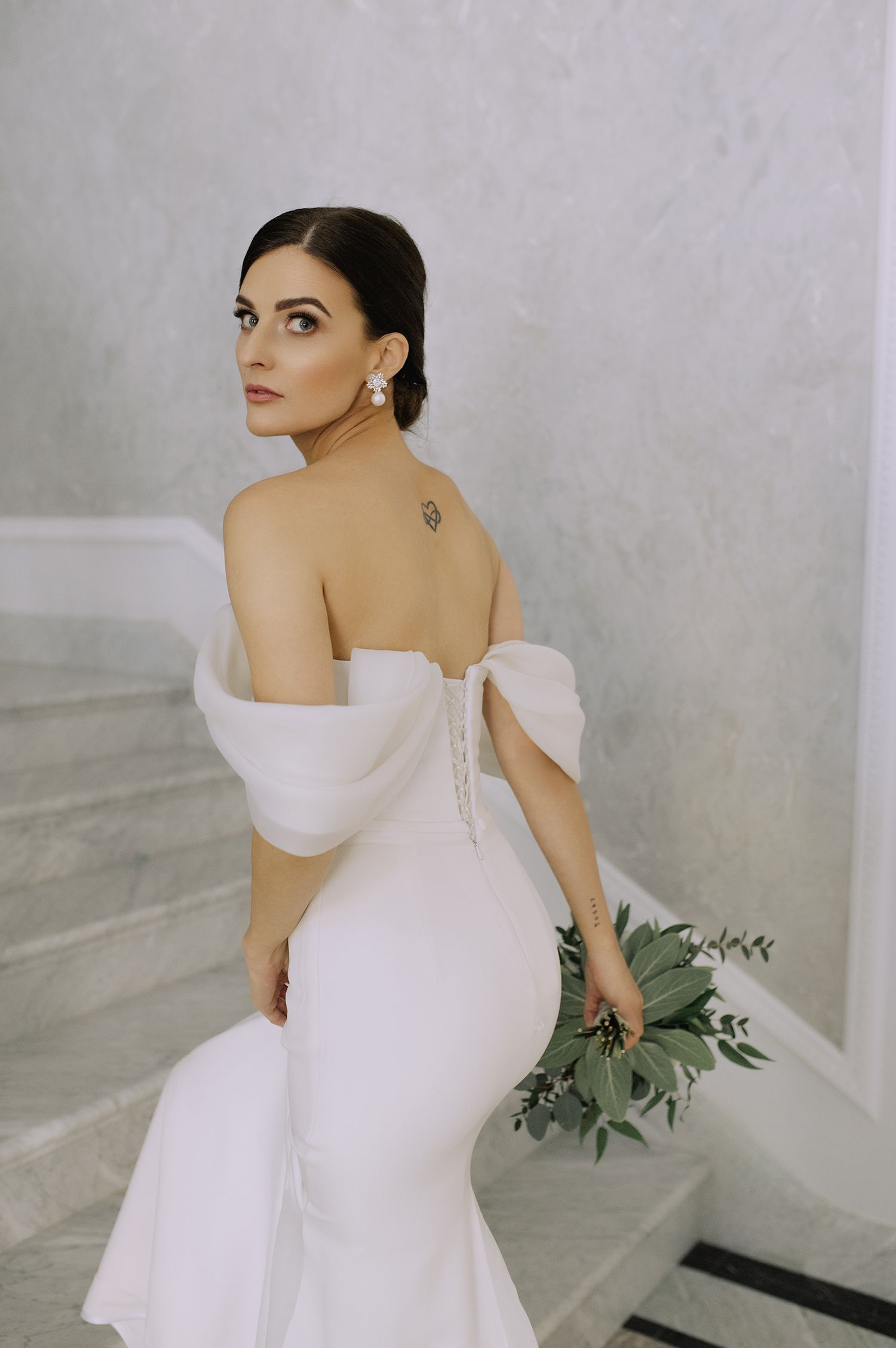 Elegant bride in a sleek off-shoulder white wedding dress poses against a light marble backdrop with delicate greenery.