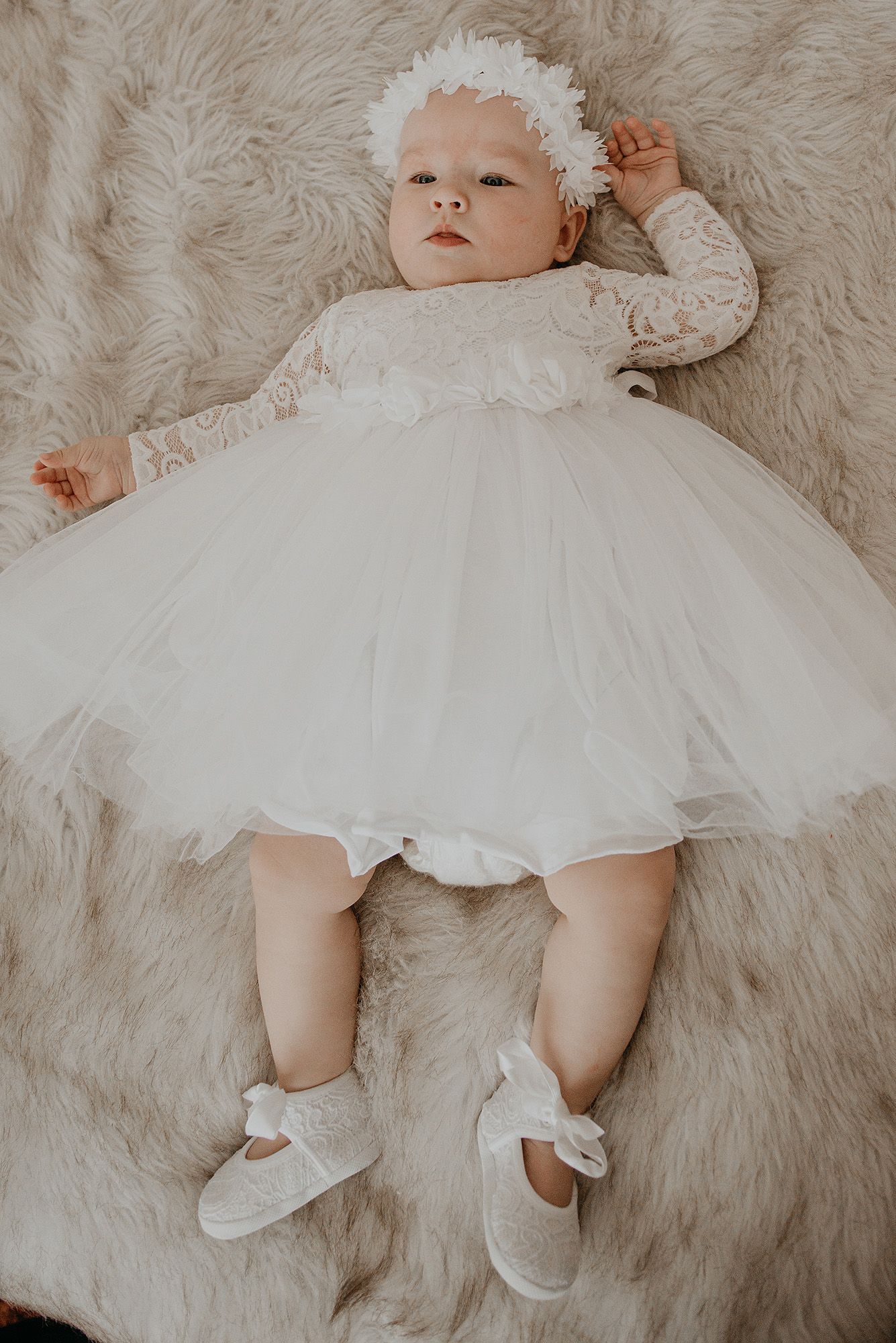 Baby wearing white dress and headband lies on plush fur rug in series of delicate portrait poses.