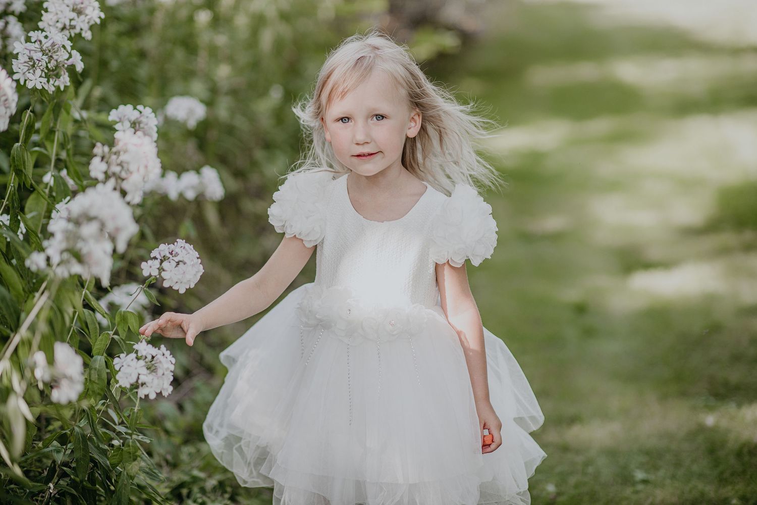 A young figure in a white tulle dress stands near blooming white flowers in a dreamy garden setting.