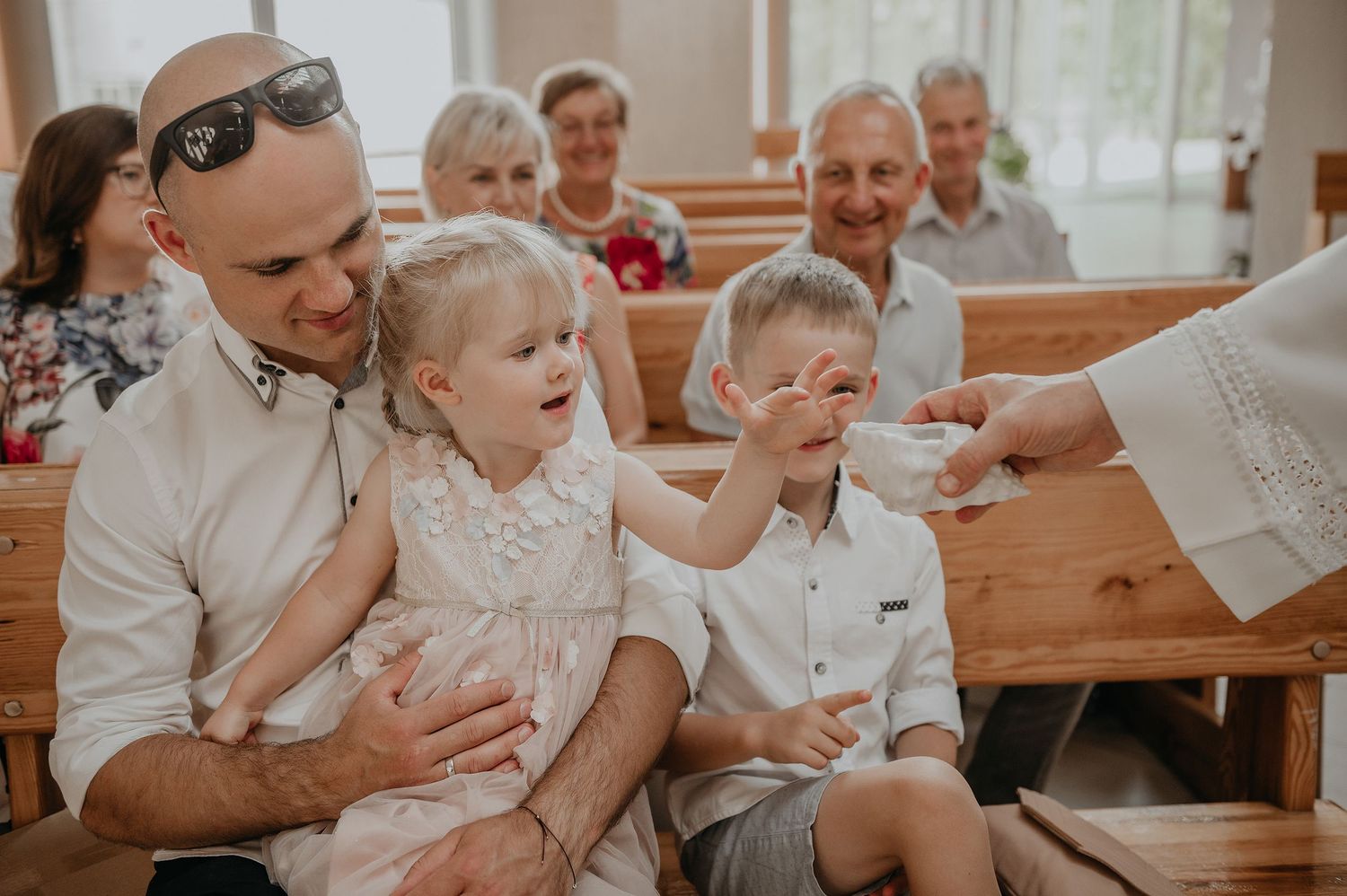 A joyful moment during a church ceremony as children reach out to touch a priest's hand in a sunlit chapel.