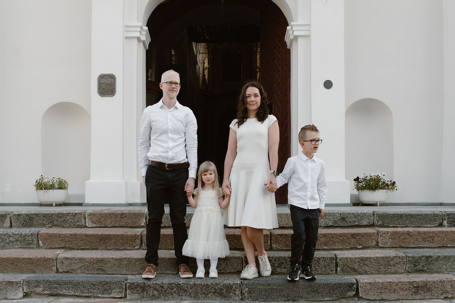 A family of four stands together on church steps wearing coordinated white and formal attire.