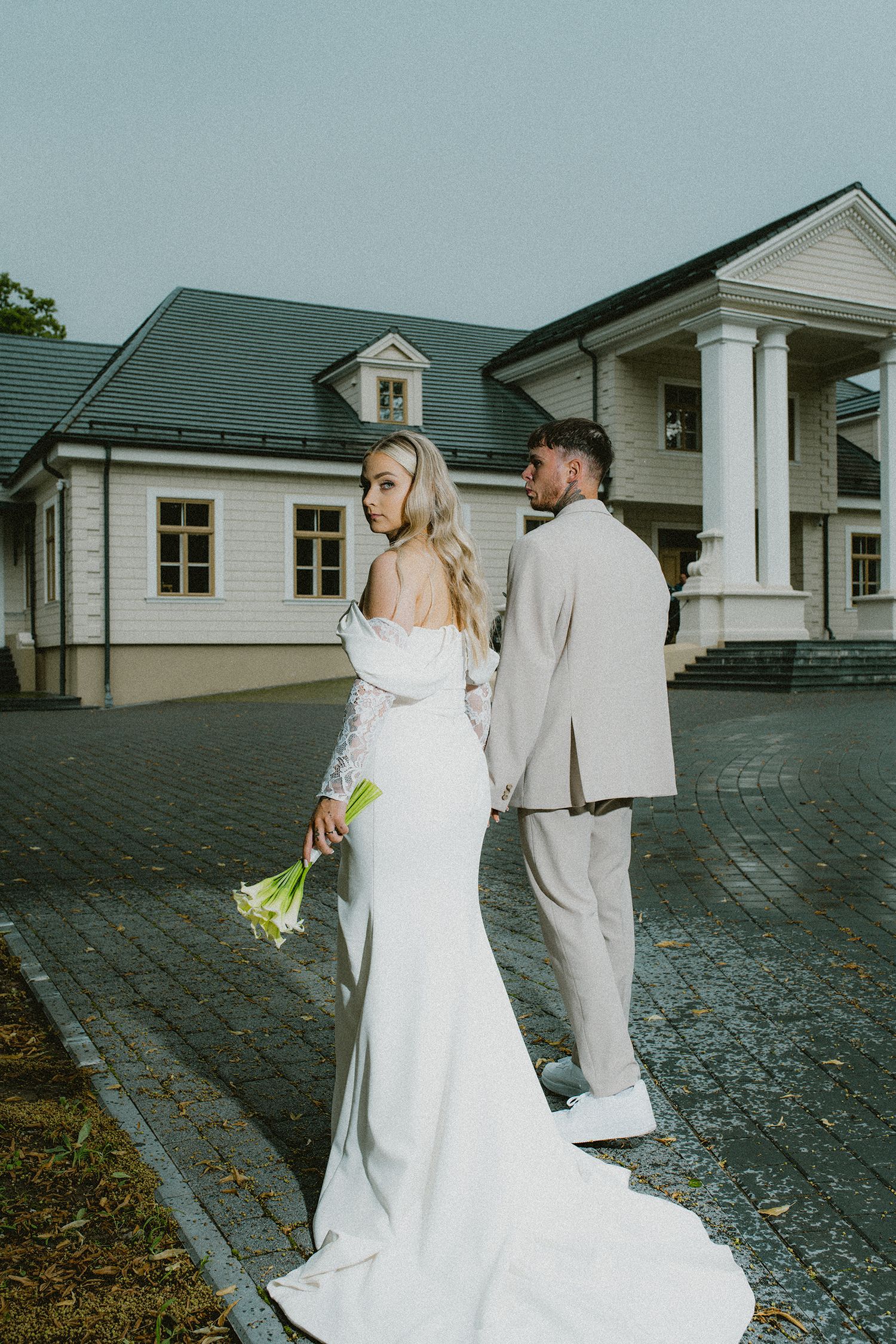 A couple in wedding attire walk together on a cobblestone path near white colonial buildings.
