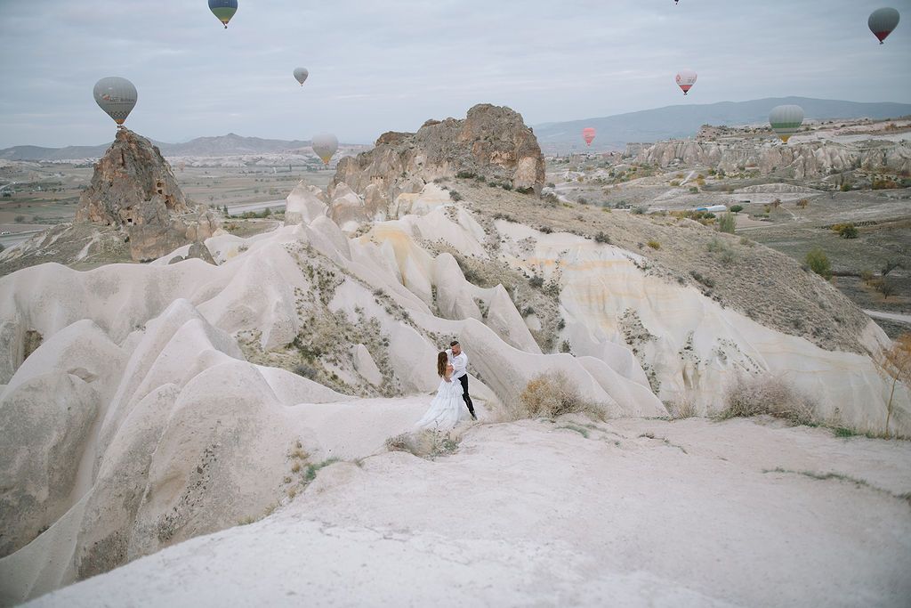 Couple in wedding attire poses on white rock formations in Cappadocia with hot air balloons floating above.