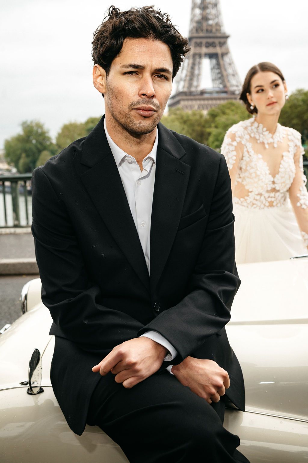 Man in black suit sits on white car with Eiffel Tower and bride in lace gown in background, Paris.