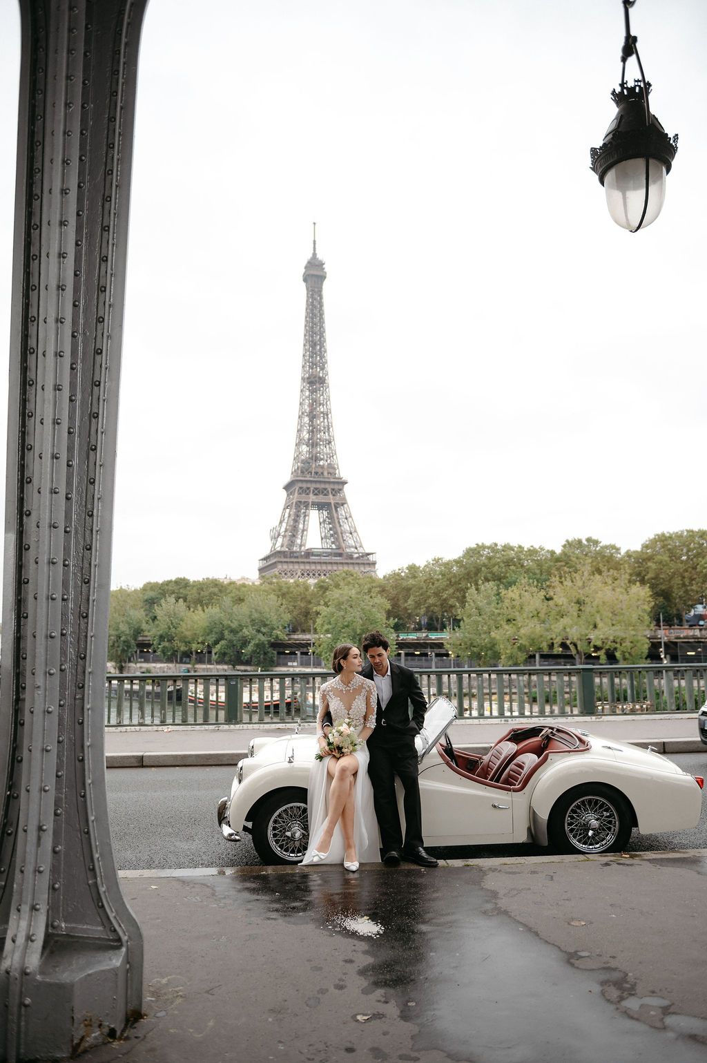Couple poses with vintage white convertible car on wet Parisian bridge with Eiffel Tower backdrop.