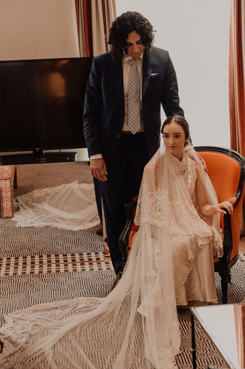 A groom in a navy suit adjusts his bride's lace veil as she sits in an orange chair wearing a white wedding gown.