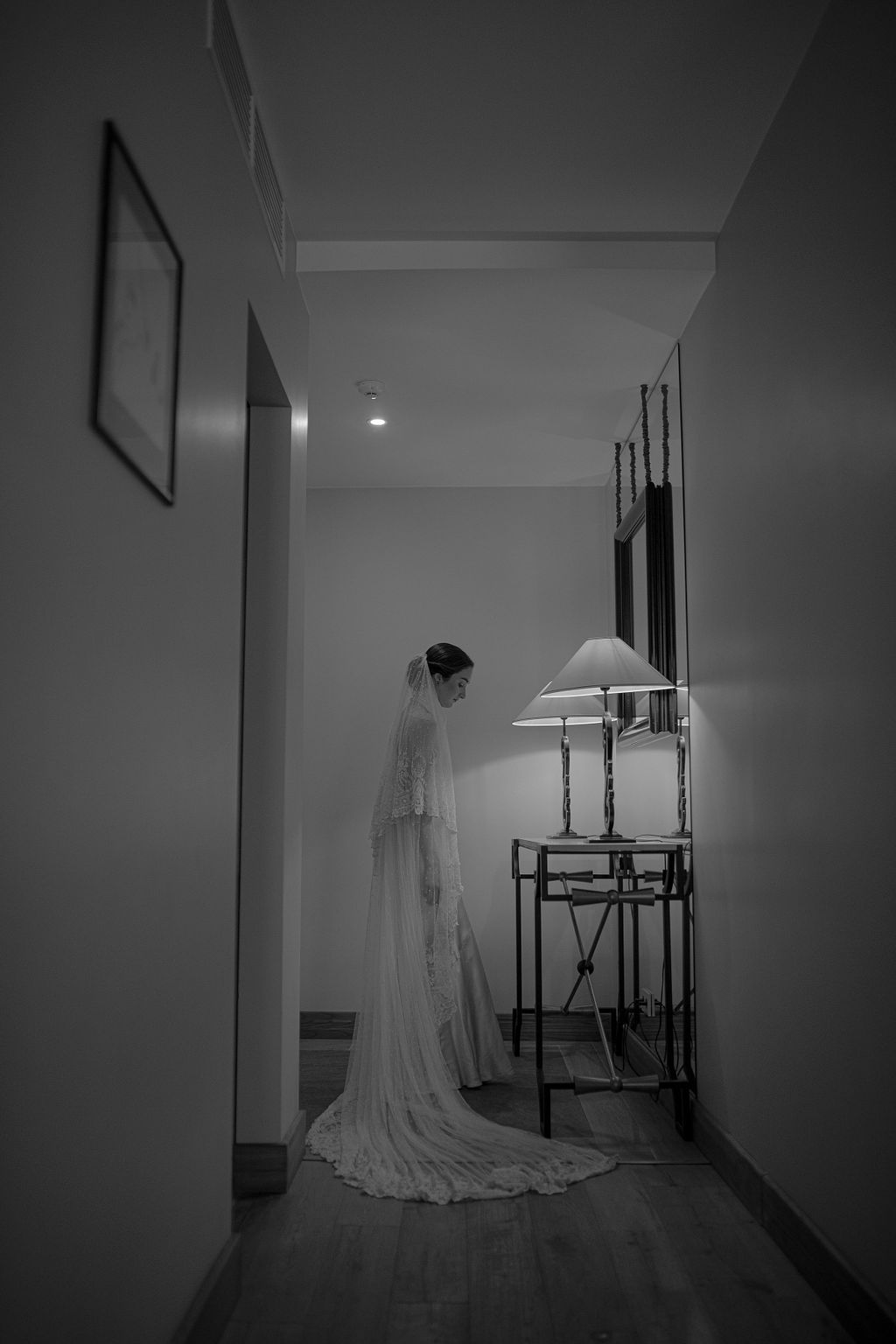 Black and white photo of a bride in a flowing wedding gown and veil standing in a hallway beside a lit lamp.