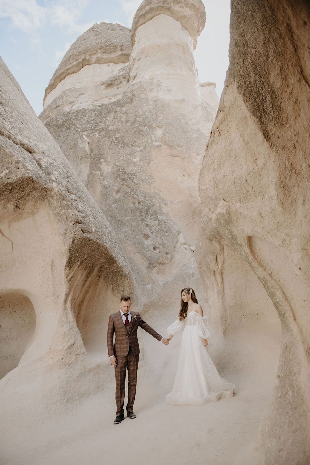 Bride and groom hold hands among towering fairy chimneys in Cappadocia, Turkey, wearing elegant wedding attire.