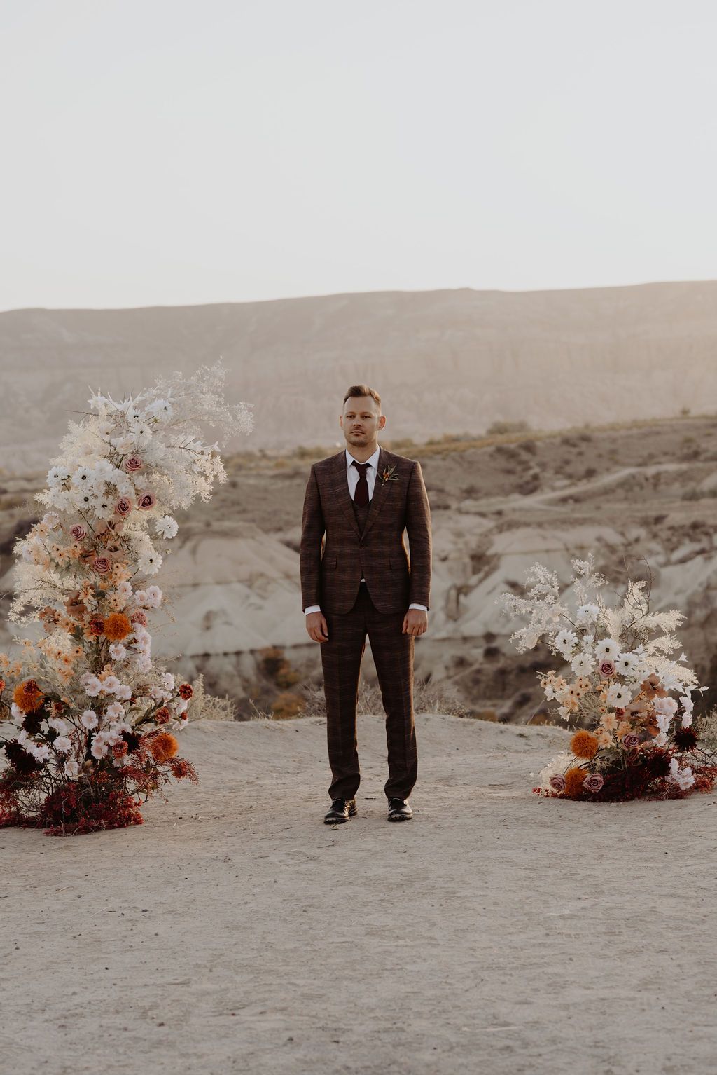Groom in dark plaid suit standing between floral arrangements in a desert landscape.